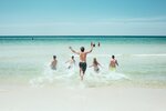 Das Bild zeigt Sonnenschein und blauen Himmel. Eine Gruppe junger Menschen läuft ins Meer hinein. Wasser spritzt auf.